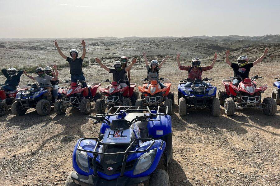 Un groupe de touristes joyeux levant les bras en signe de victoire lors d'une excursion en quad dans le désert d'Agafay. Les participants portent des casques et sont alignés sur leurs quads colorés (bleus, rouges et oranges) face au vaste paysage aride et rocailleux sous un ciel clair.